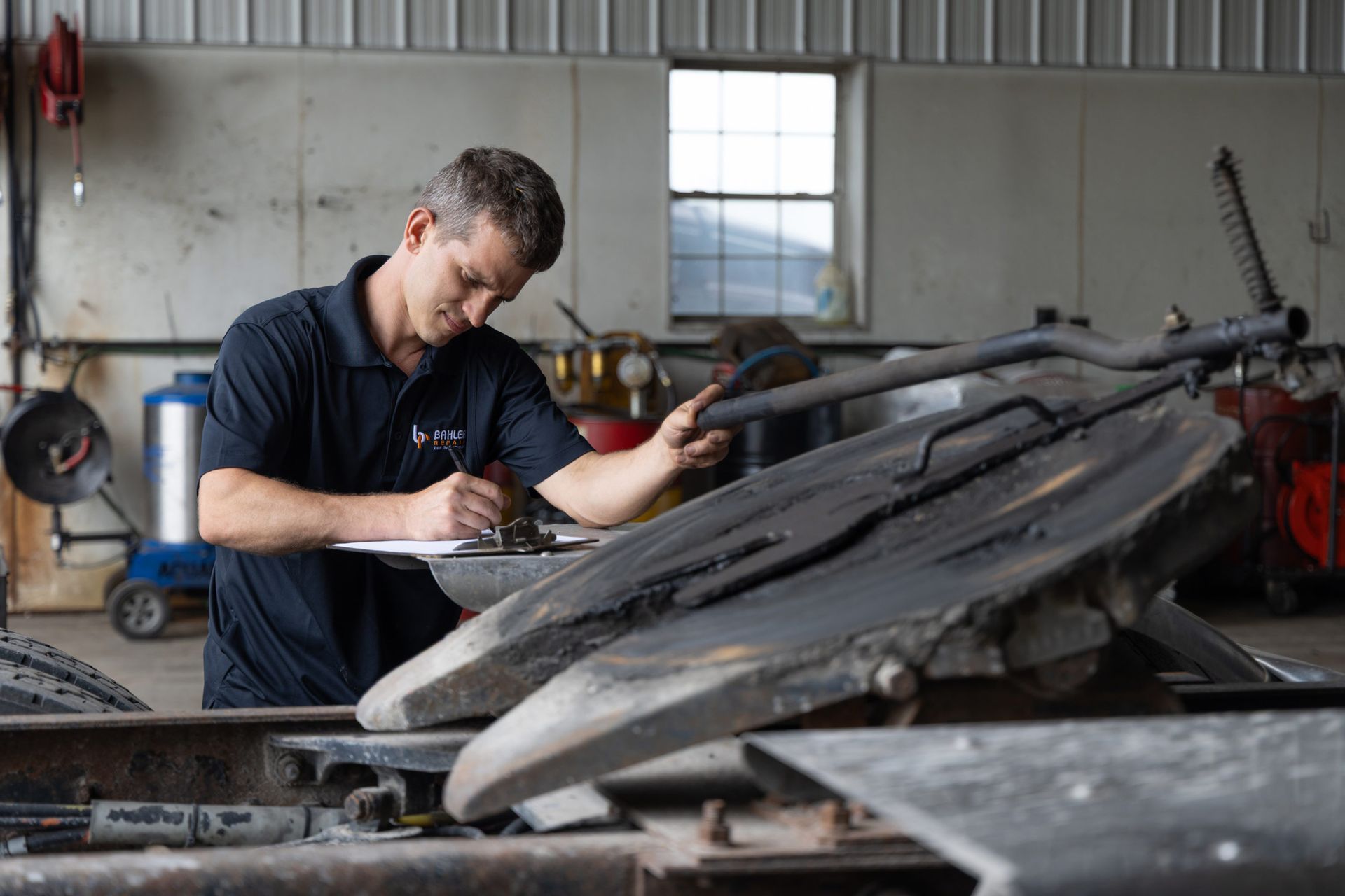 Mechanic inspecting a truck's fifth wheel hitch, writing on a clipboard in a garage setting.