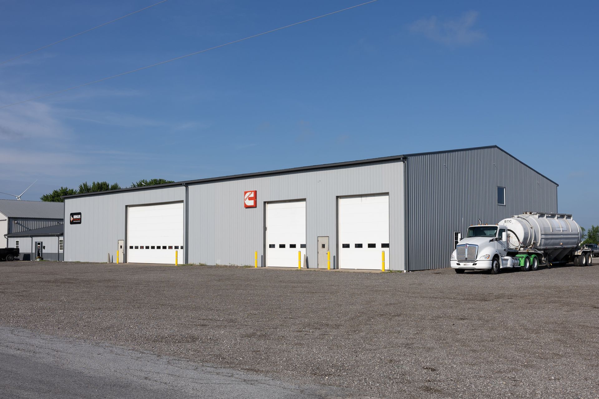 A gray industrial building with three garage doors, a tanker truck parked outside, and a blue sky.