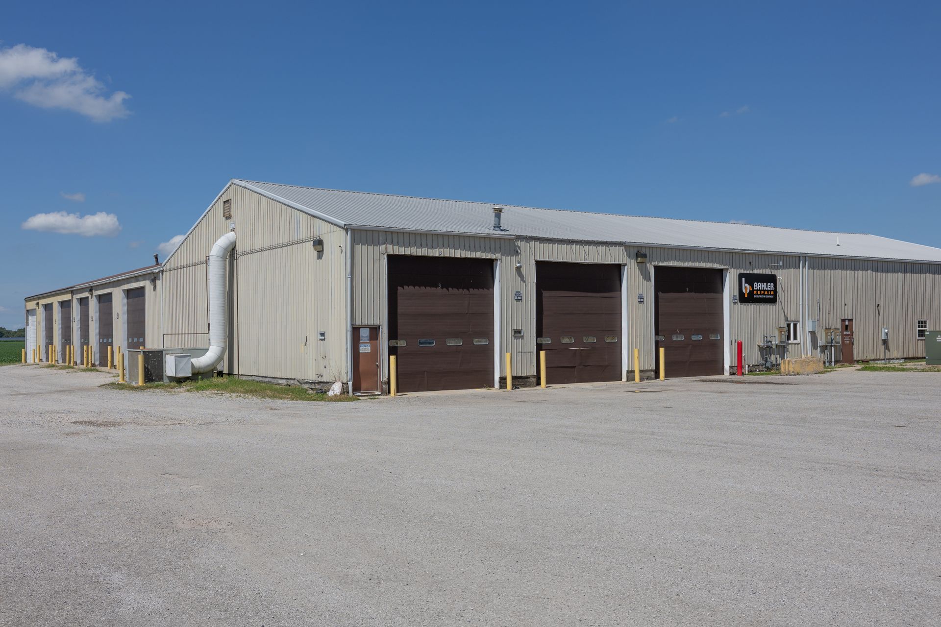 Gray industrial building with multiple garage doors under a clear blue sky. Gravel lot in the foreground.