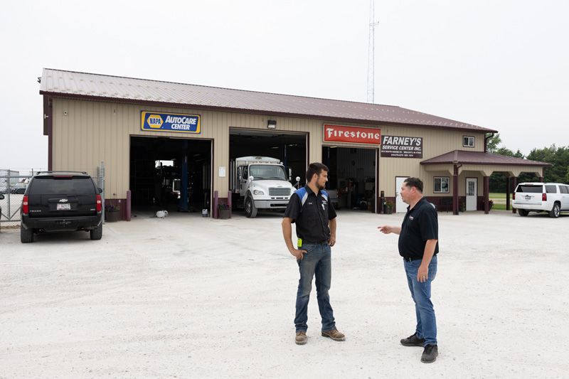 Two men converse outside a car repair shop, which has a garage door open, revealing a van inside. The setting is outdoors on a gravel lot.