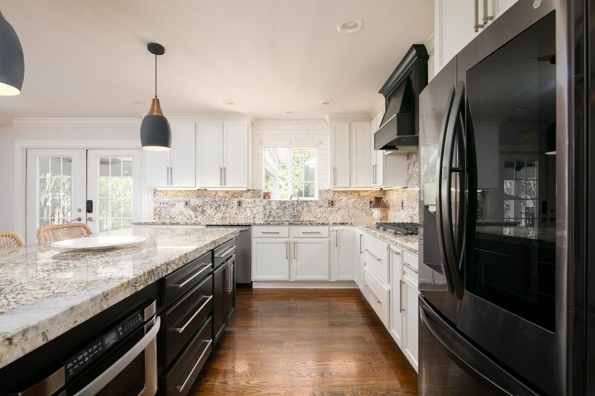 A modern kitchen featuring white cabinets, dark base cabinets, granite countertops, a black range hood, and wood floors.