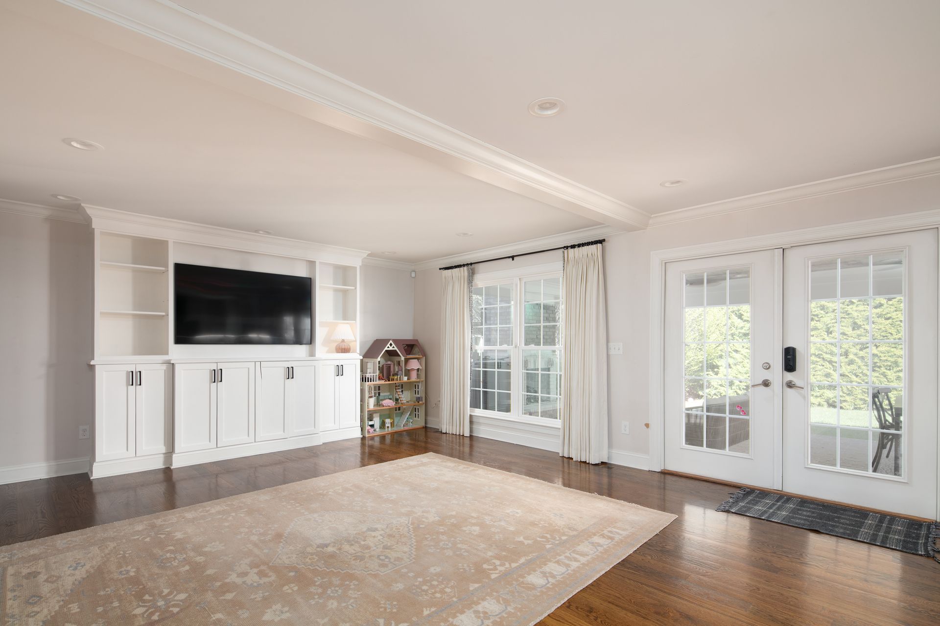 A bright, minimalist living room with a large wall unit, built-in TV, wood floors, a beige rug, and a double glass door.