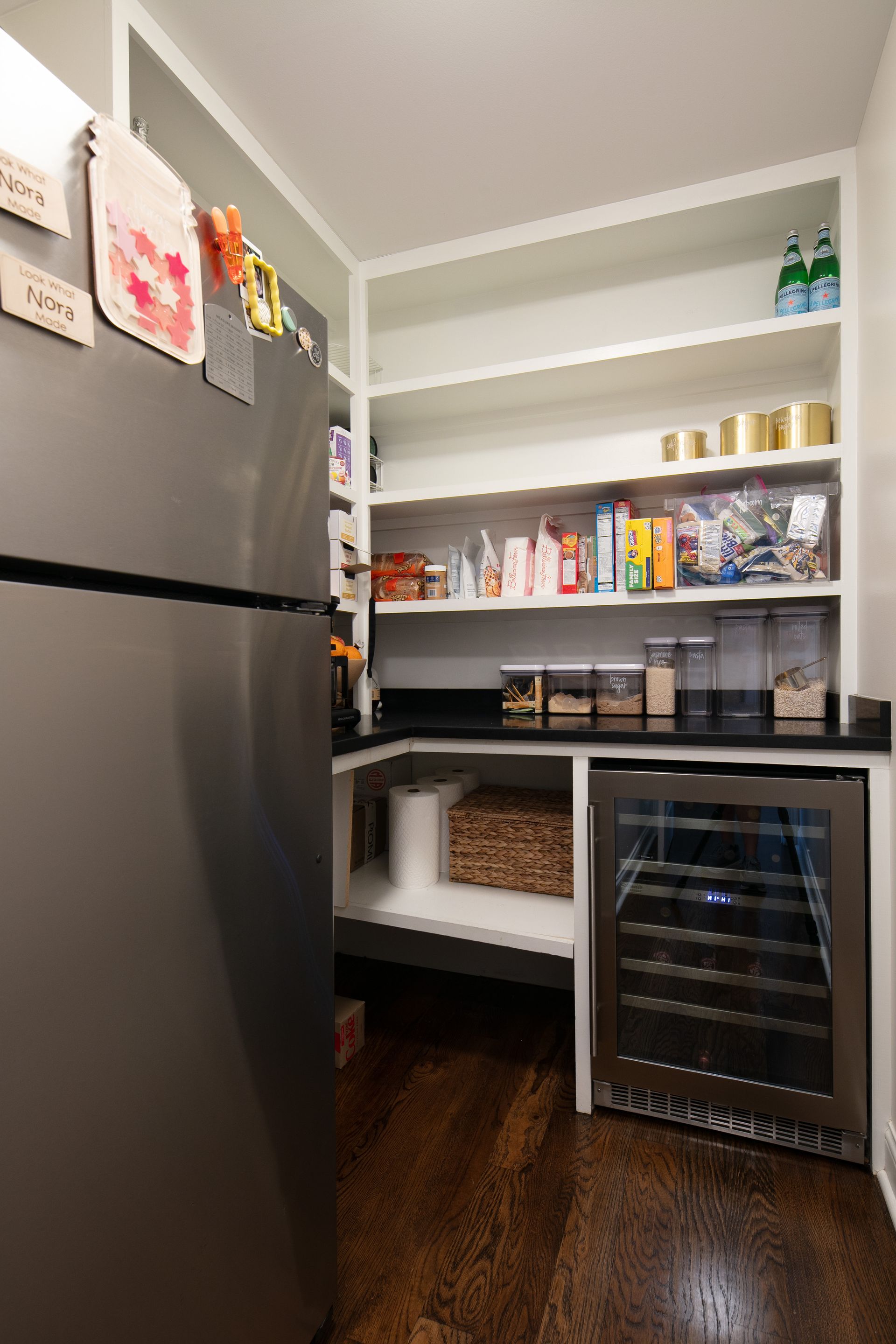 A kitchen pantry featuring a dark stainless refrigerator, open shelving with food items, a wine fridge, and wood floors.