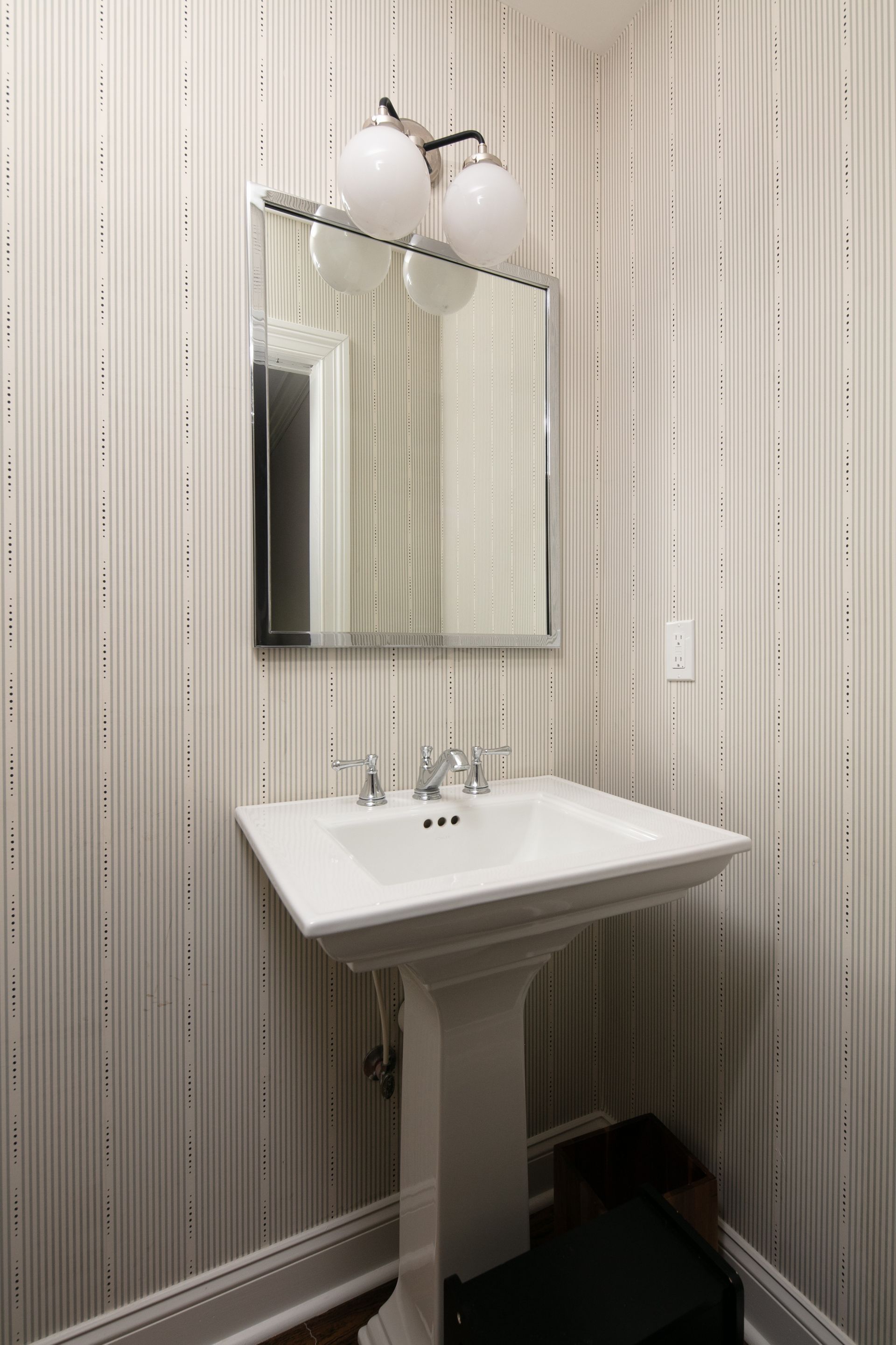 A pedestal sink and a rectangular mirror mounted on a wall covered in vertical-striped patterned wallpaper.