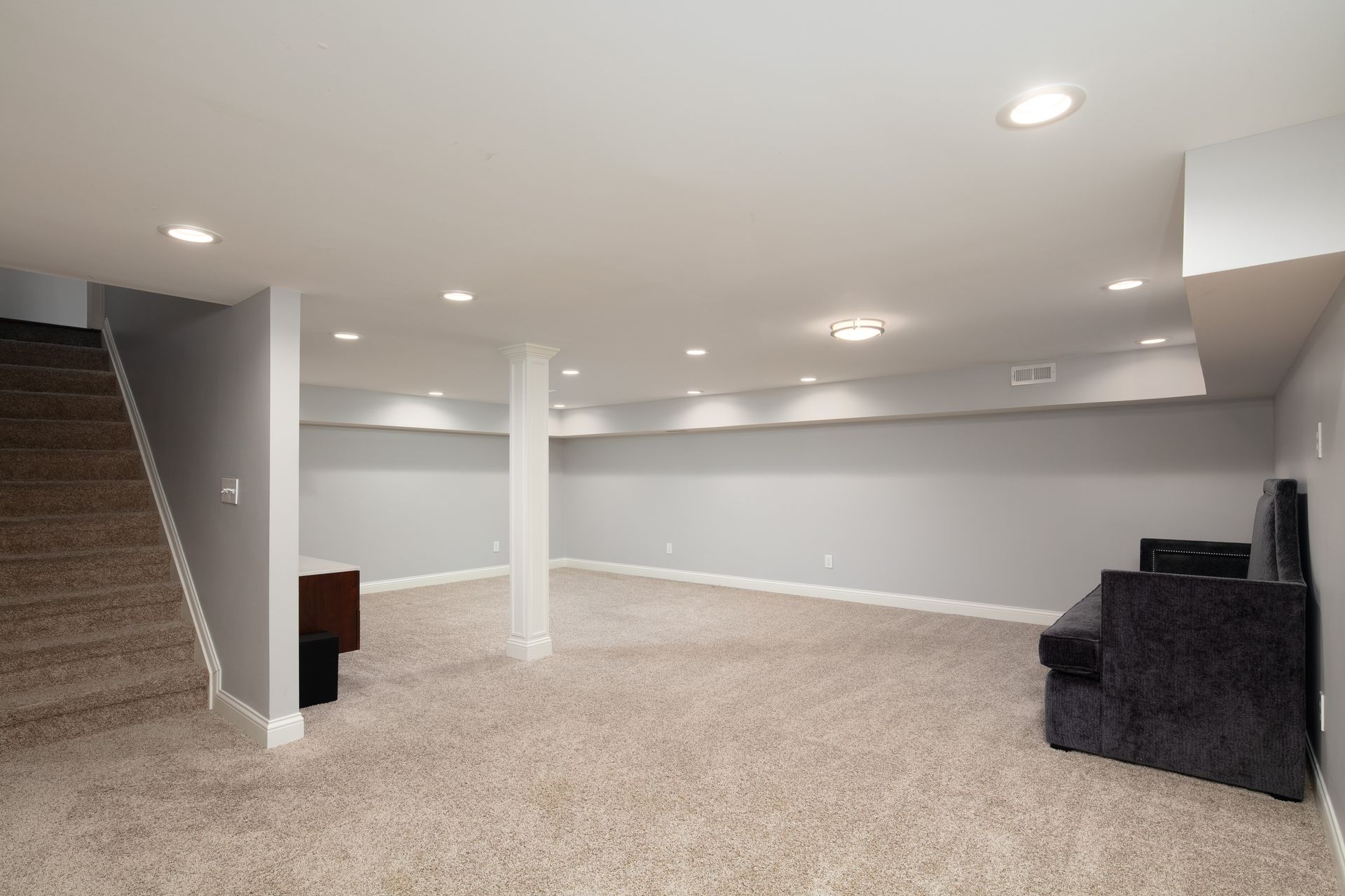 A finished basement with gray walls, light-colored carpet, a central white support pillar, and a dark sofa.
