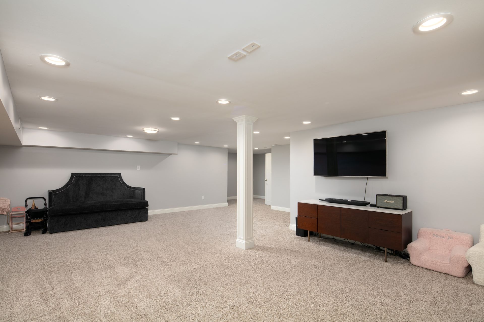 A finished basement with light gray walls, carpet, a central support column, a wall-mounted TV, and assorted furniture.