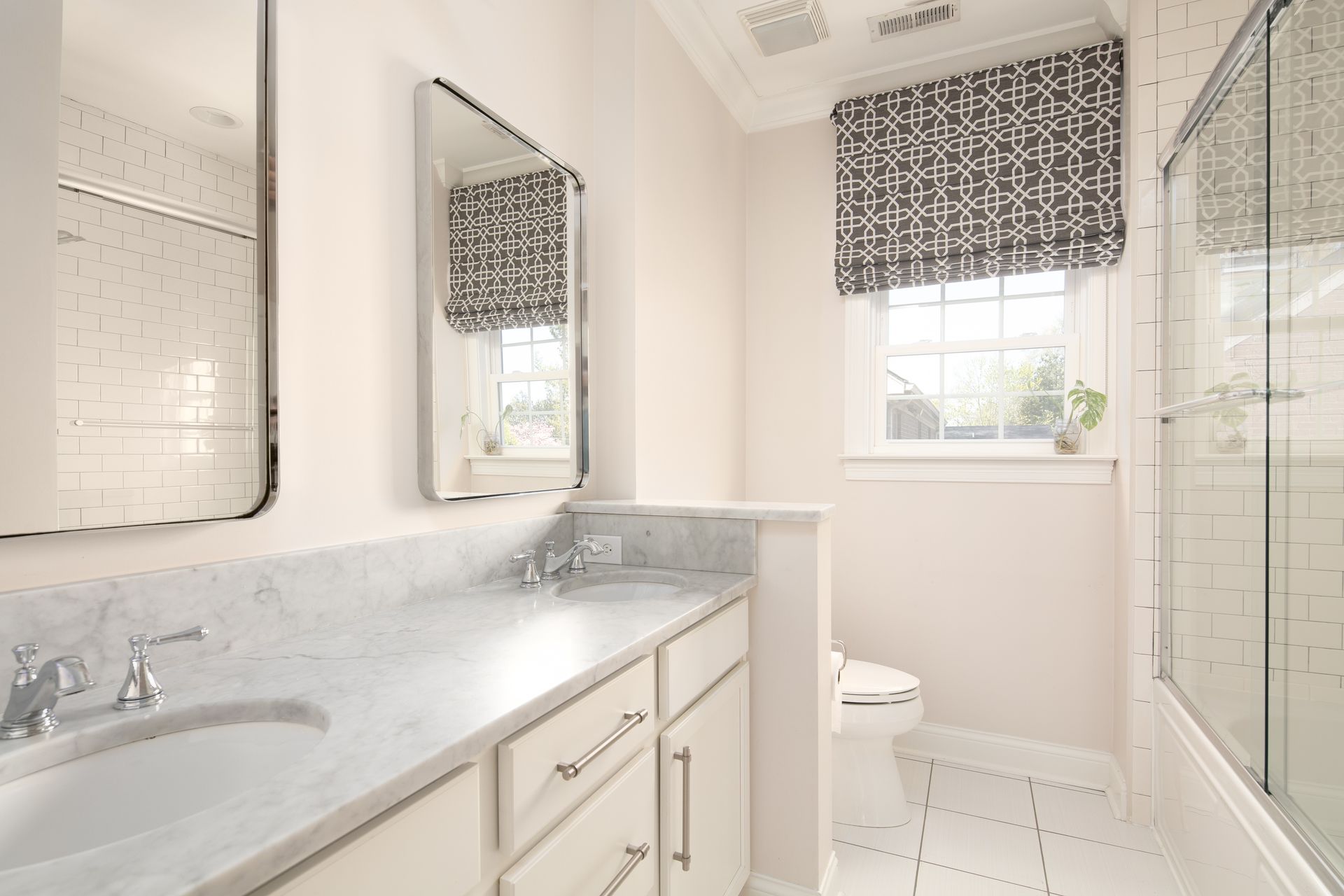 A bright, modern bathroom featuring a marble-topped vanity, dual mirrors, a toilet, and a glass-enclosed shower.