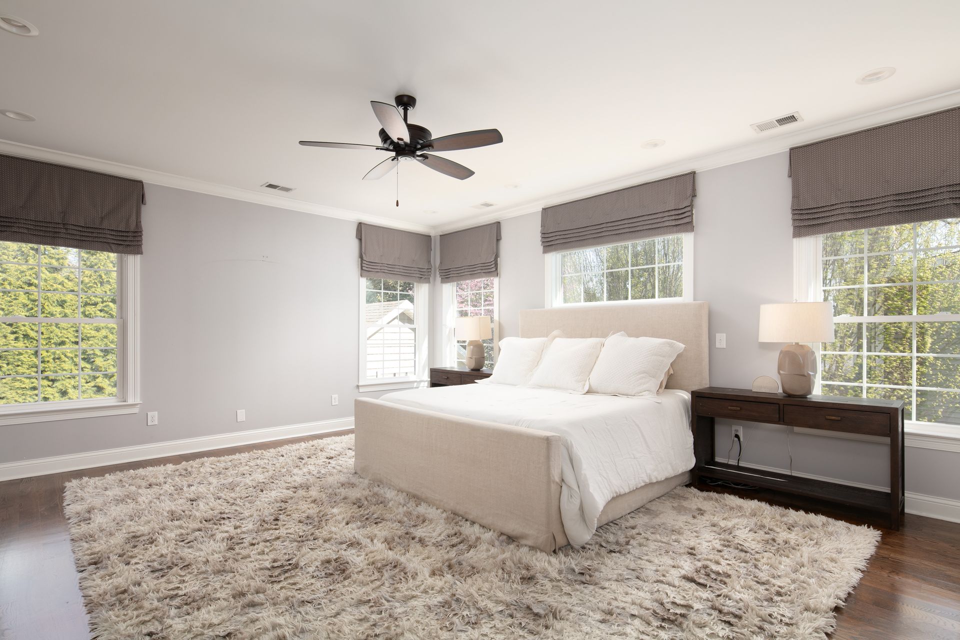 A light-colored bedroom featuring a large bed, a plush area rug, windows with striped valances, and a ceiling fan.