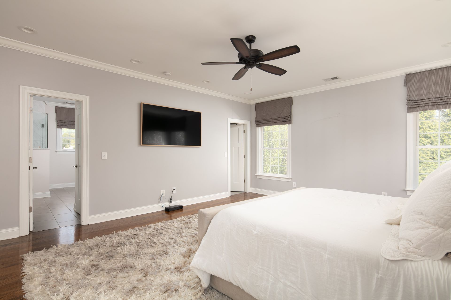 A spacious, light-grey bedroom with a large bed, fluffy white rug, wall-mounted television, and ceiling fan.