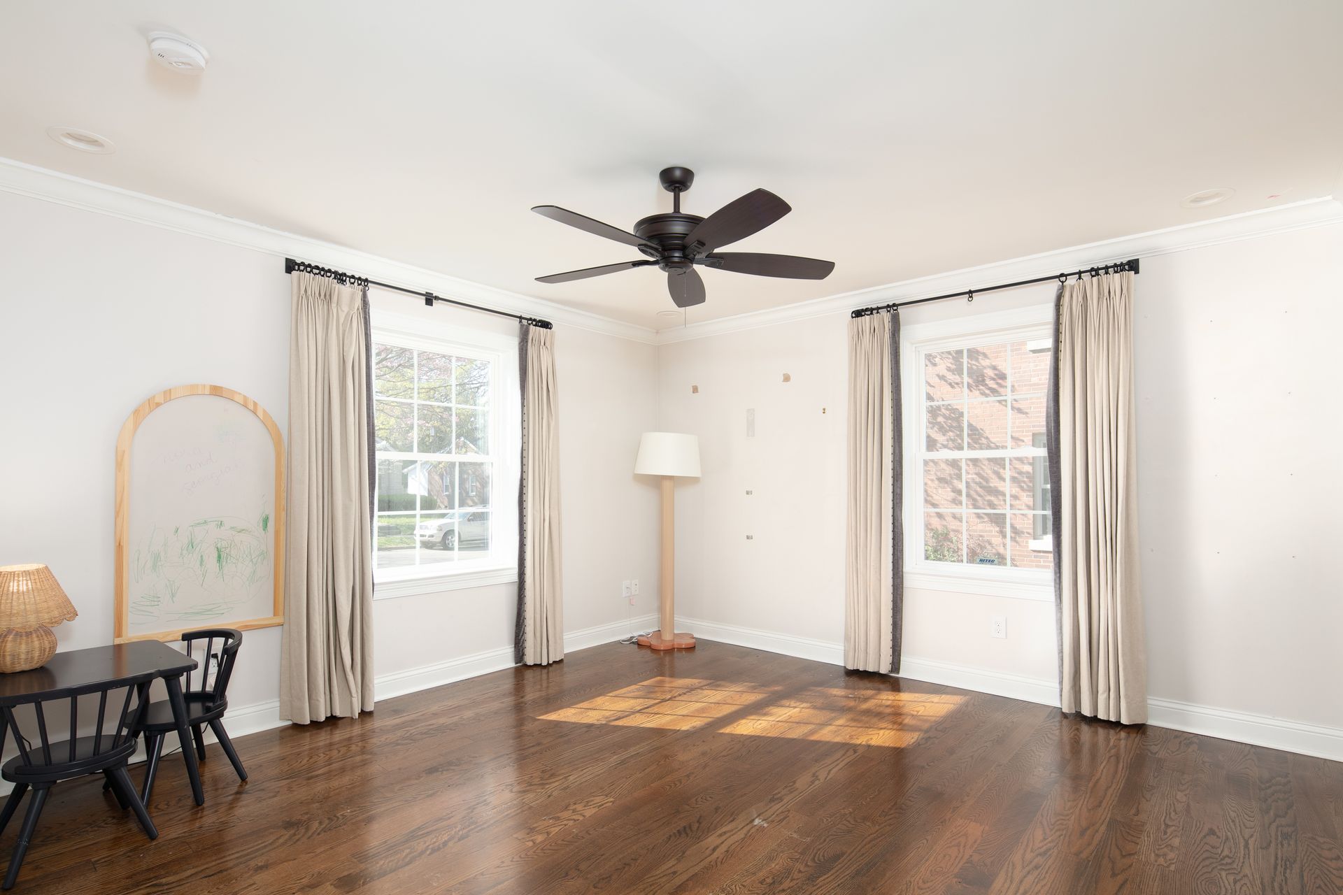 A bright, spacious room with hardwood floors, a dark ceiling fan, two windows with beige curtains, and a small desk.