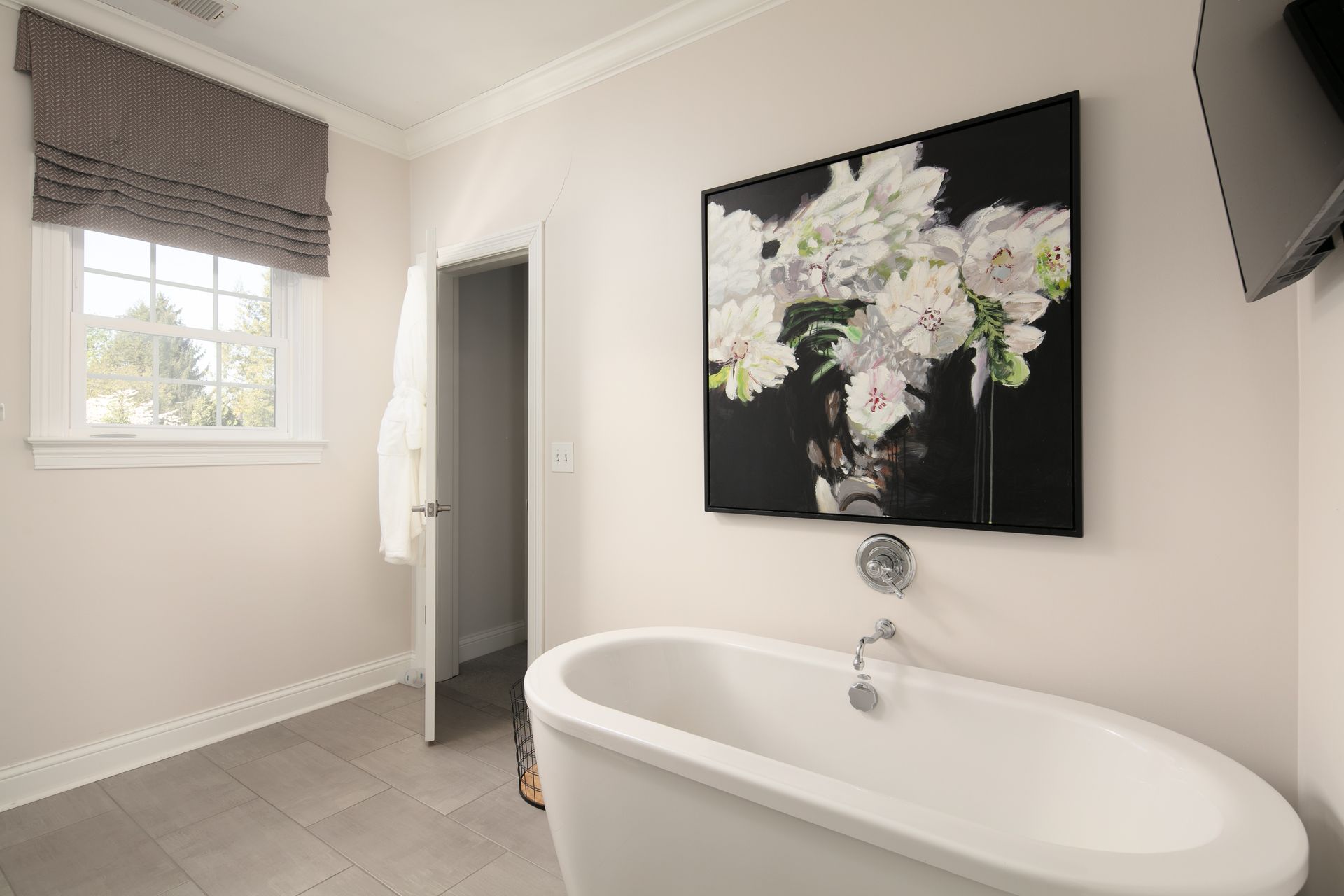 A white freestanding bathtub under a large floral painting on a beige wall, with a nearby window and an open doorway.