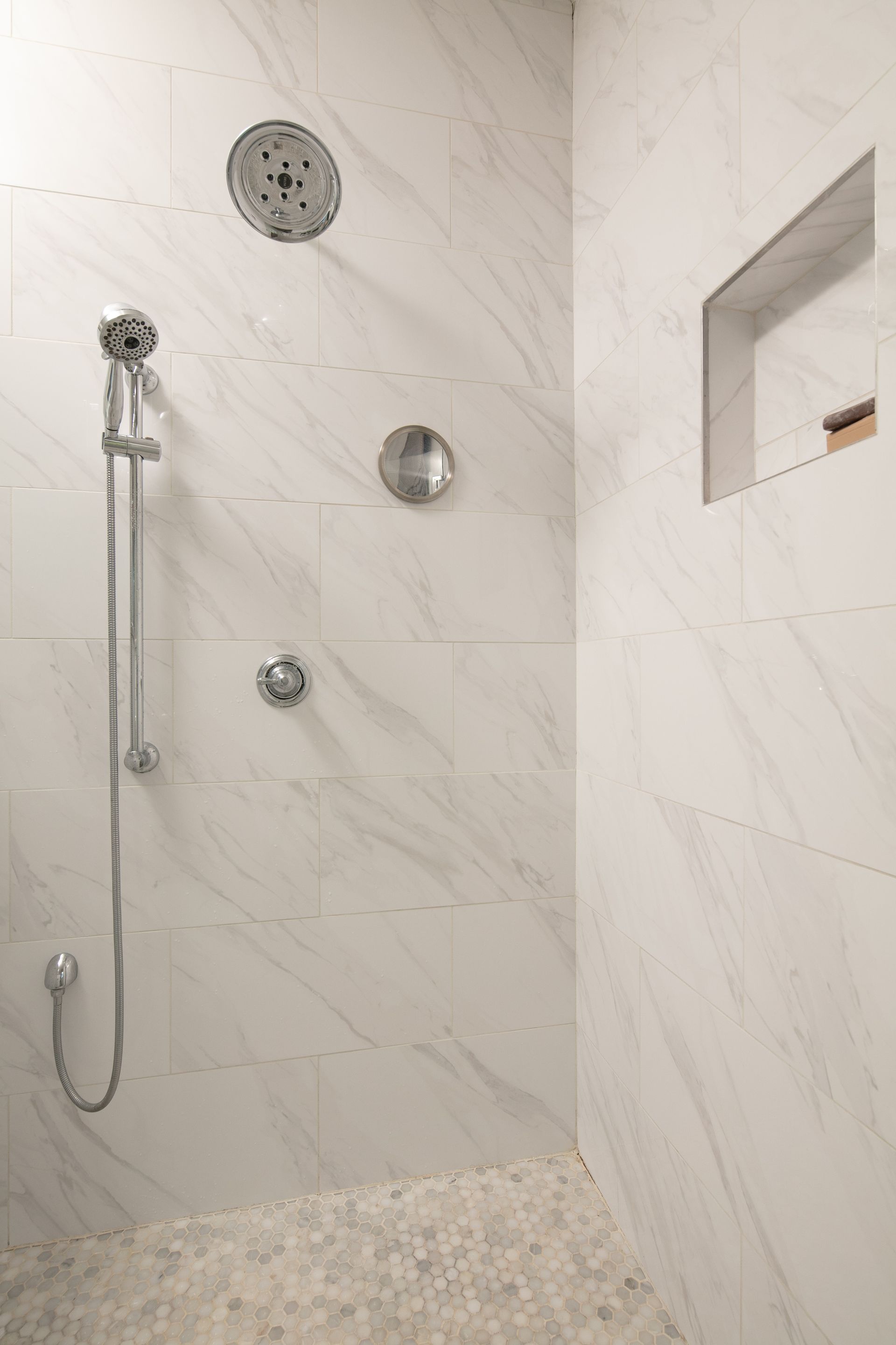 A modern shower stall featuring white marble-look tiles, a rainfall showerhead, handheld sprayer, and built-in wall niche.