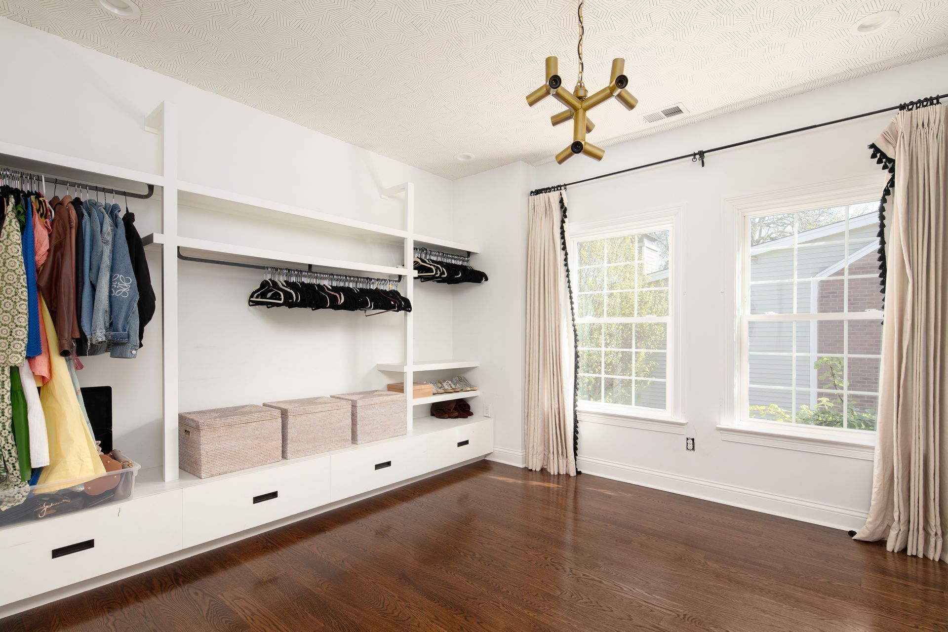 A white walk-in closet with open shelving, hanging clothes, storage bins, wood floors, and two windows with curtains.