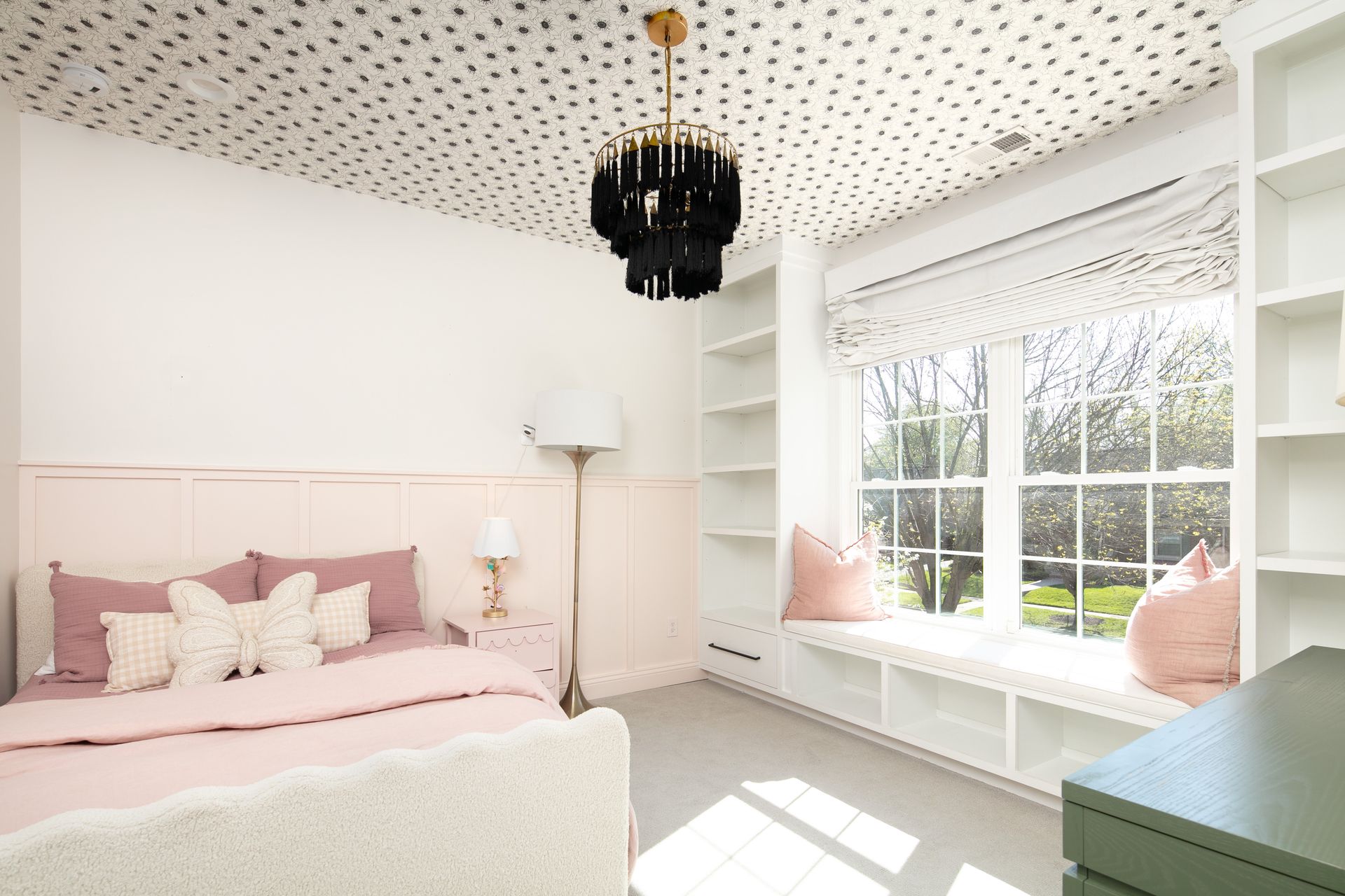 A bedroom features a bed with blush bedding, a dotted ceiling, a black chandelier, and a window seat with built-in shelves.