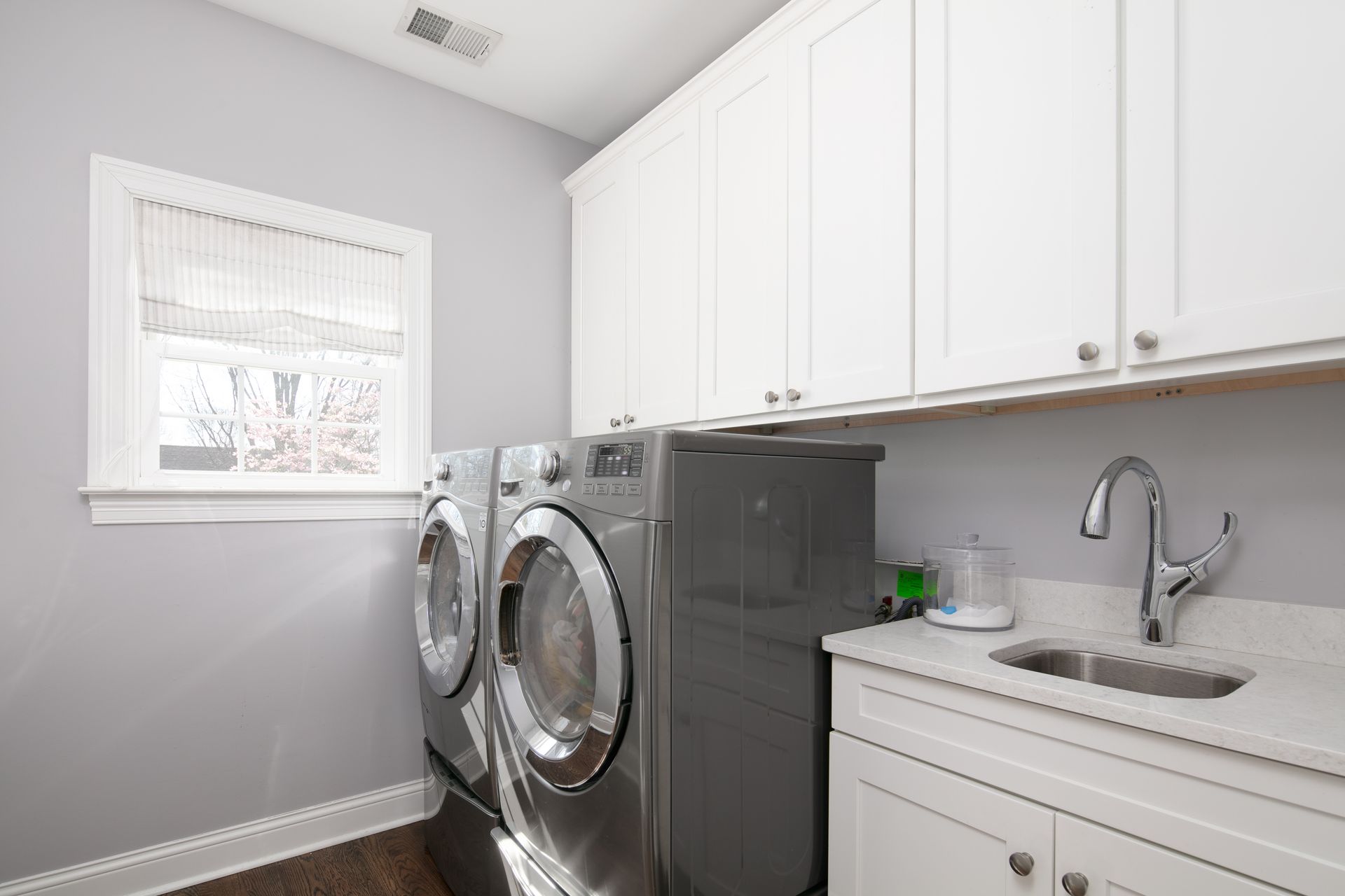 A bright laundry room with gray walls, white cabinets, a sink, and two side-by-side gray washing machines.