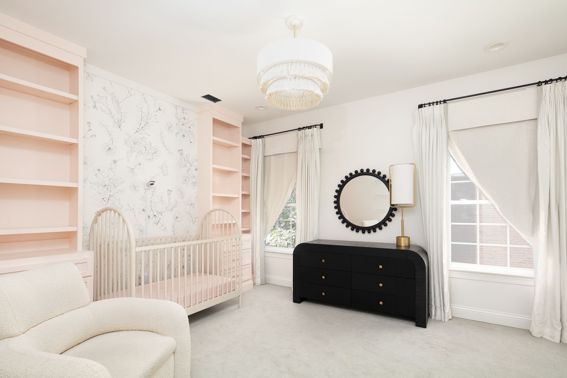 A nursery featuring pink shelving, a white crib, a black dresser with a round mirror, and a beige armchair.