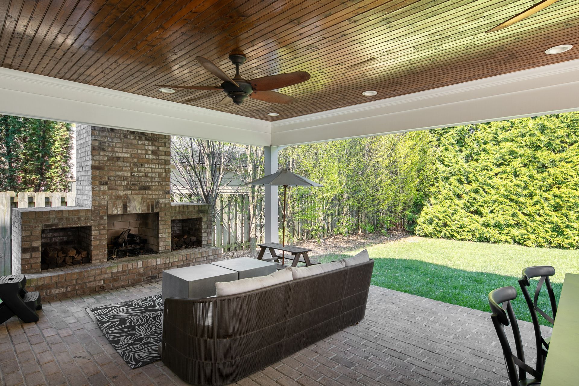 A spacious outdoor covered patio features a stone fireplace, wicker furniture, and a ceiling fan, overlooking a yard.