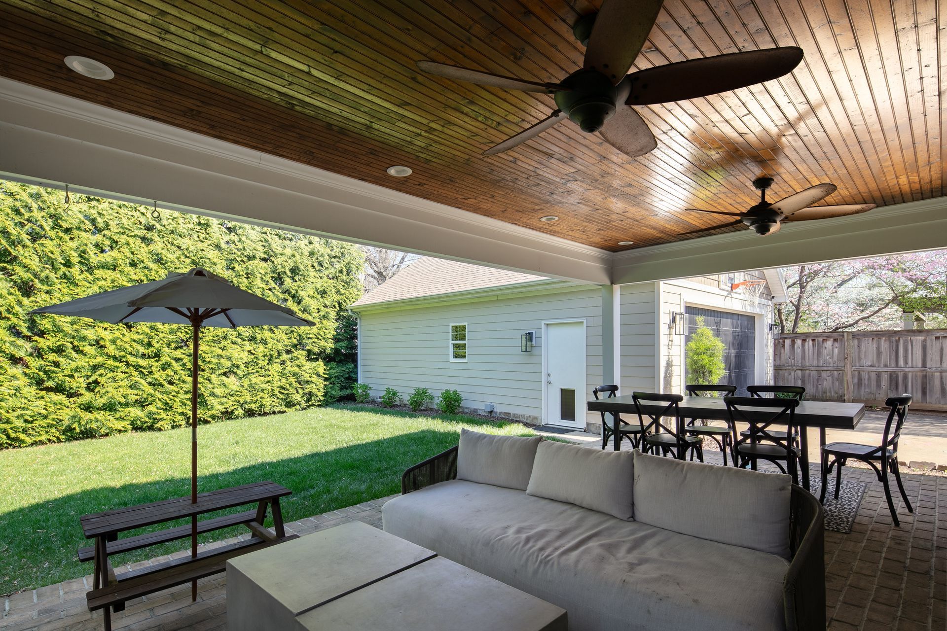 A covered patio with a wooden ceiling, ceiling fans, light-colored sofa, dining table, and a green yard with an umbrella.