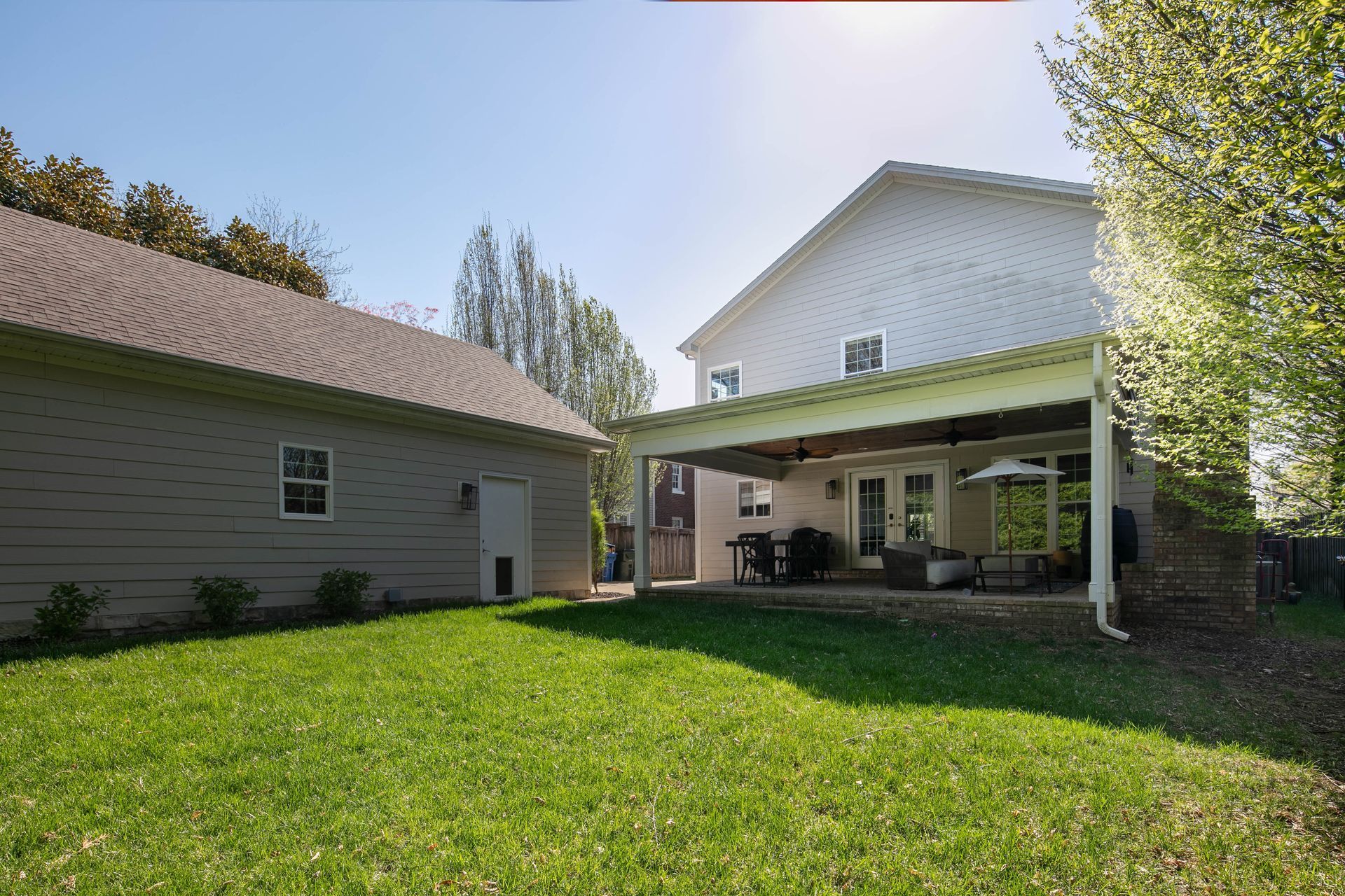 A backyard view of a light-colored two-story house with a patio roof and an adjacent garage under a bright blue sky.