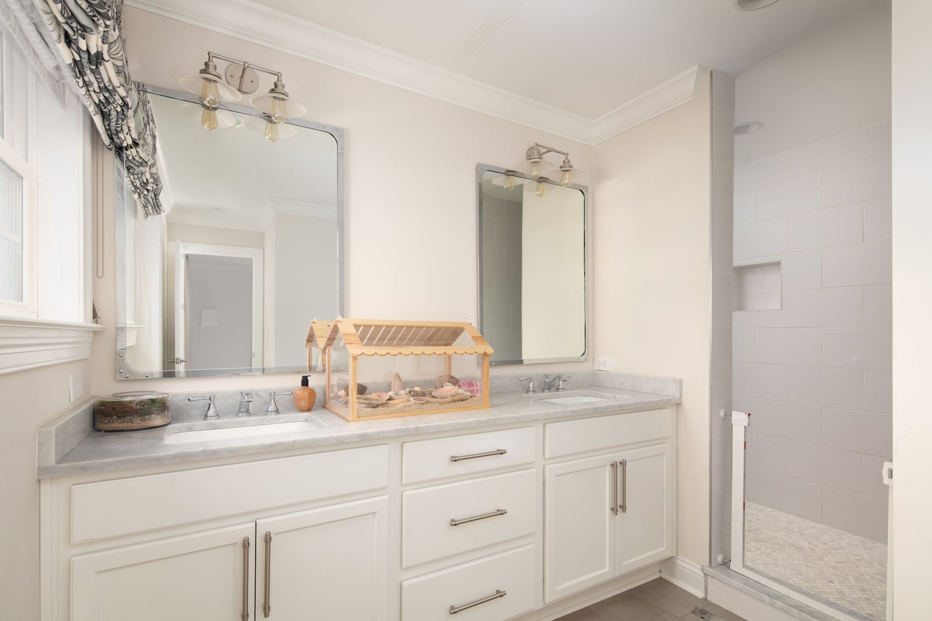 A white bathroom vanity with double sinks, two mirrors, a wooden organizer, and a glass-enclosed shower.