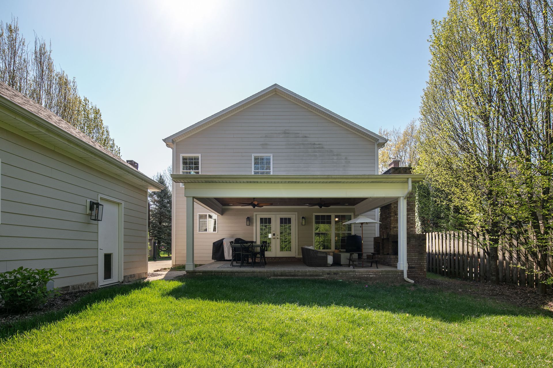 A two-story tan house with a covered back patio, outdoor furniture, and a grassy backyard on a sunny day.