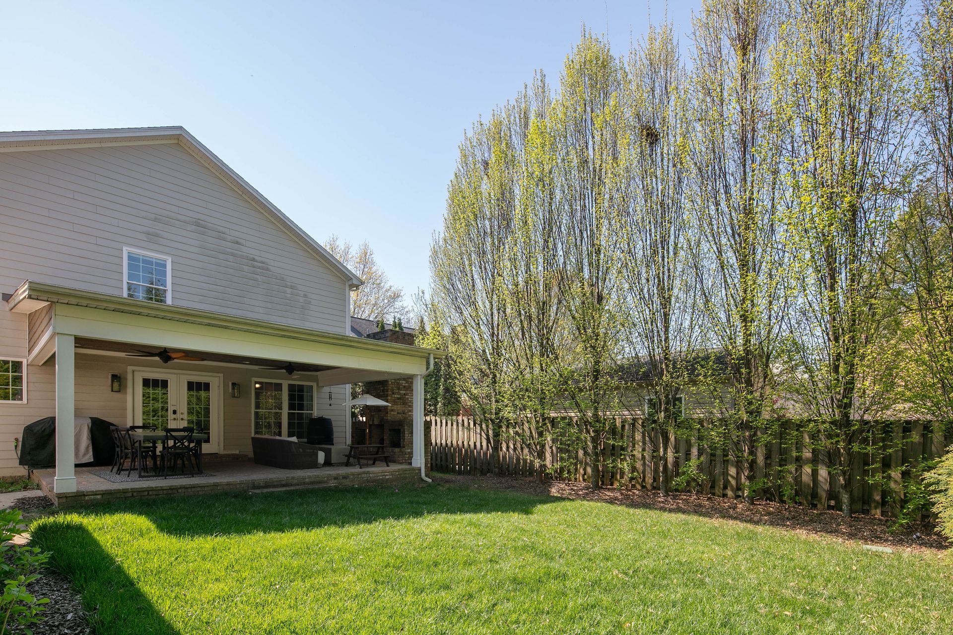 A light-colored house with a backyard patio, lawn, and a row of tall trees along a fence.
