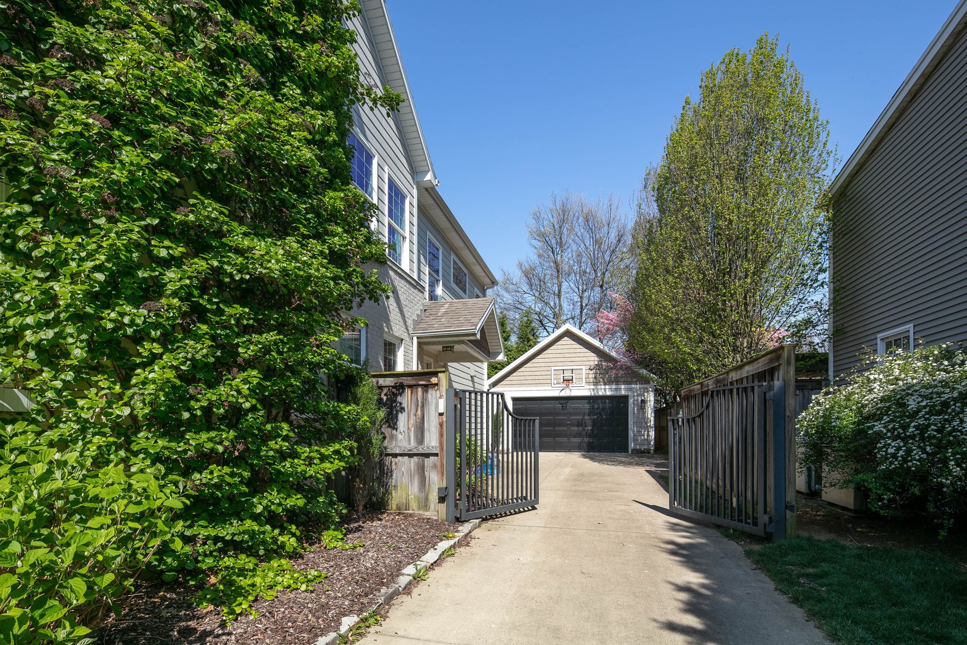 A concrete driveway leads to a detached garage between two houses, flanked by lush green trees and a metal fence.