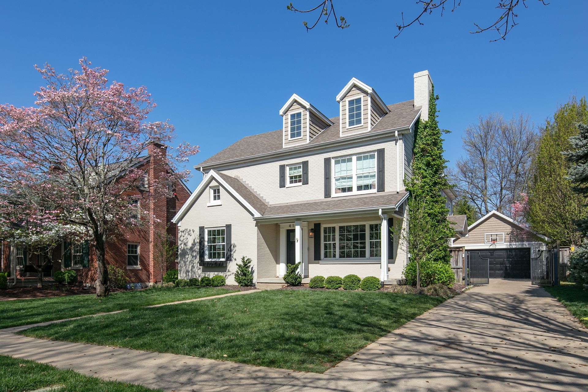 A beige, two-story house with dormer windows, a front porch, and a paved driveway under a clear blue sky.