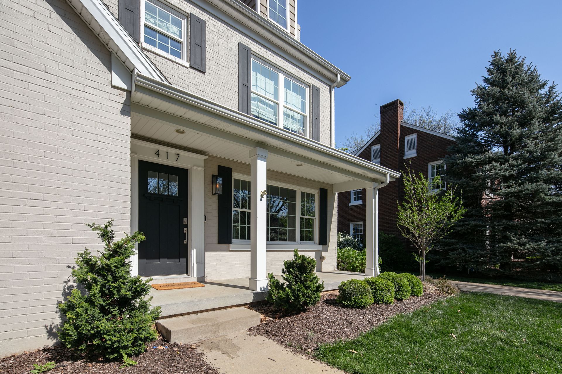 A light-colored brick house with a front porch, dark front door, and green landscaping under a clear blue sky.