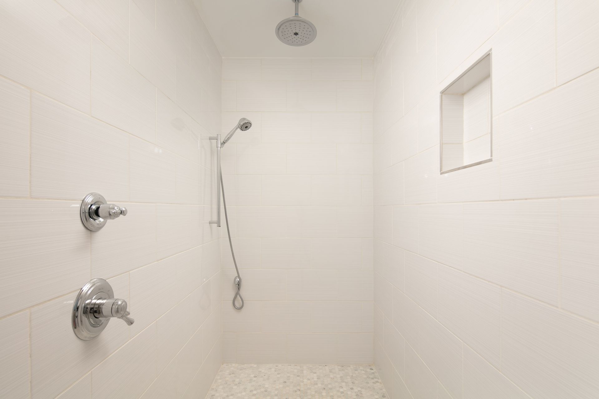 A bright white tiled shower with a rain showerhead, handheld sprayer, chrome fixtures, and a recessed wall niche.