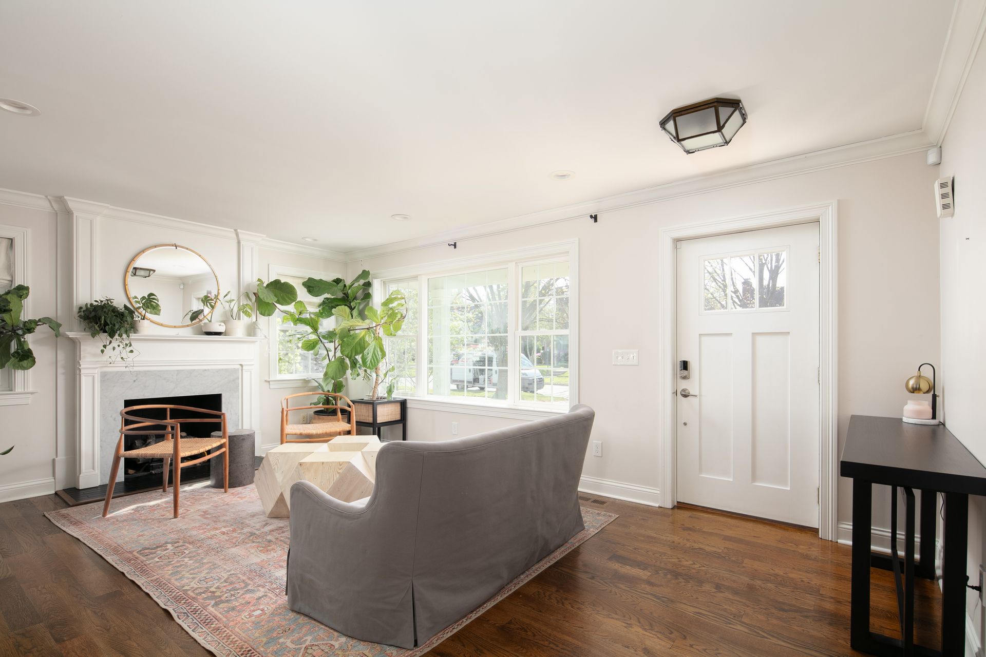 A brightly lit living room with a gray sofa, a fireplace, large windows, and a white front door on hardwood floors.