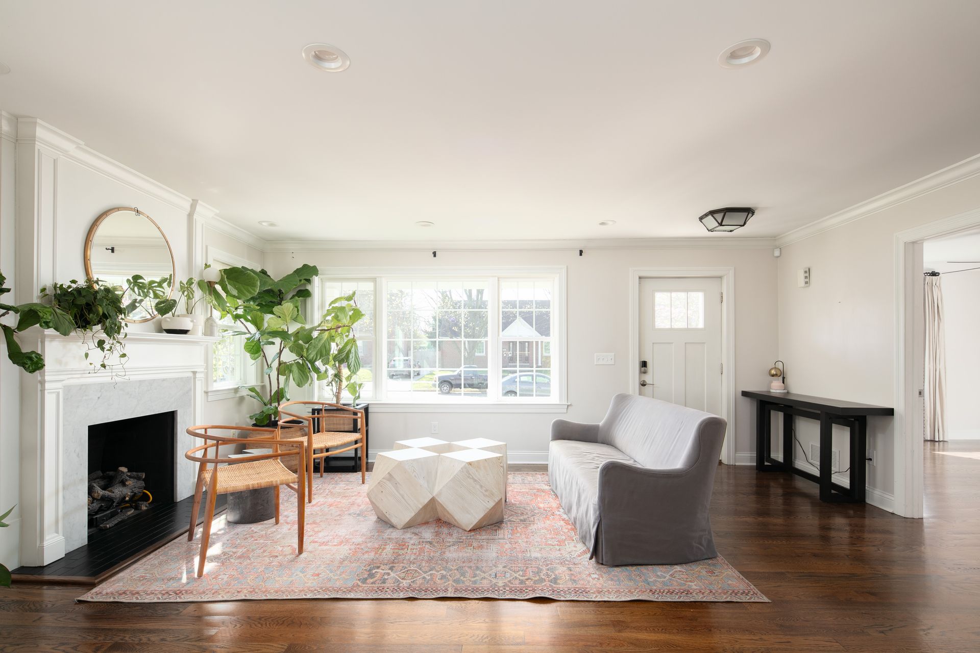 A bright, minimalist living room with hardwood floors, a fireplace, a grey sofa, a geometric coffee table, and plants.