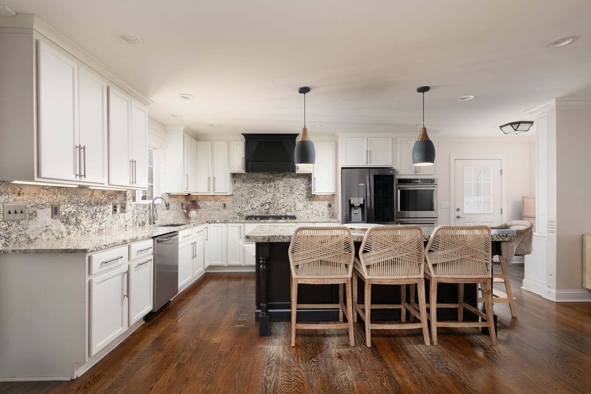 A bright, modern kitchen featuring white cabinets, granite countertops, a dark kitchen island with three wicker bar stools.