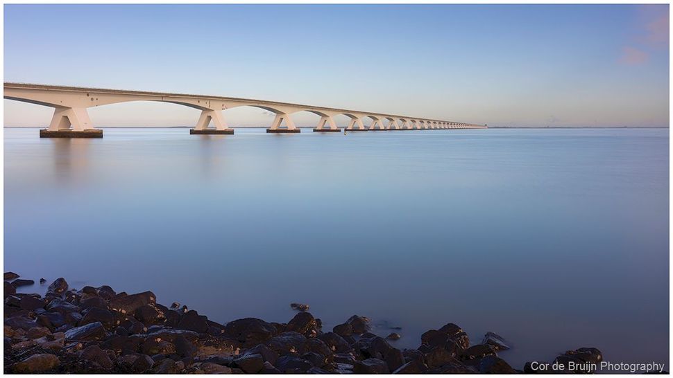 Long, arched bridge spanning calm blue water, stretching to the horizon.