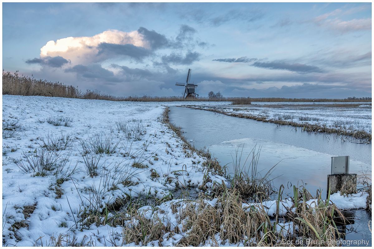 Snowy Dutch landscape with a windmill, canal, and overcast sky.