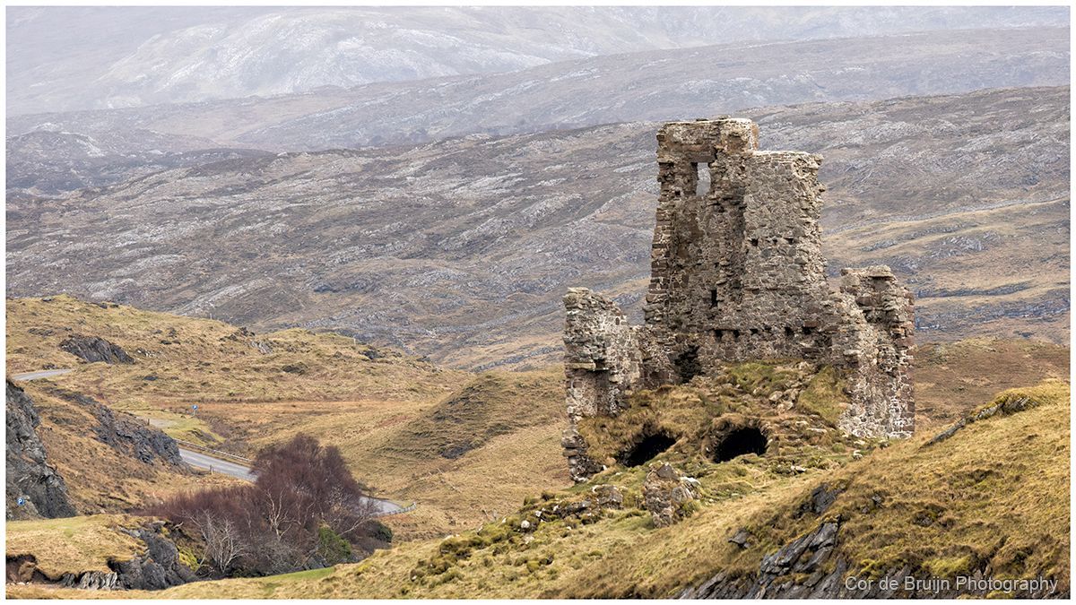 A stone ruin of an old tower stands on a grassy, hilly landscape under a cloudy sky.