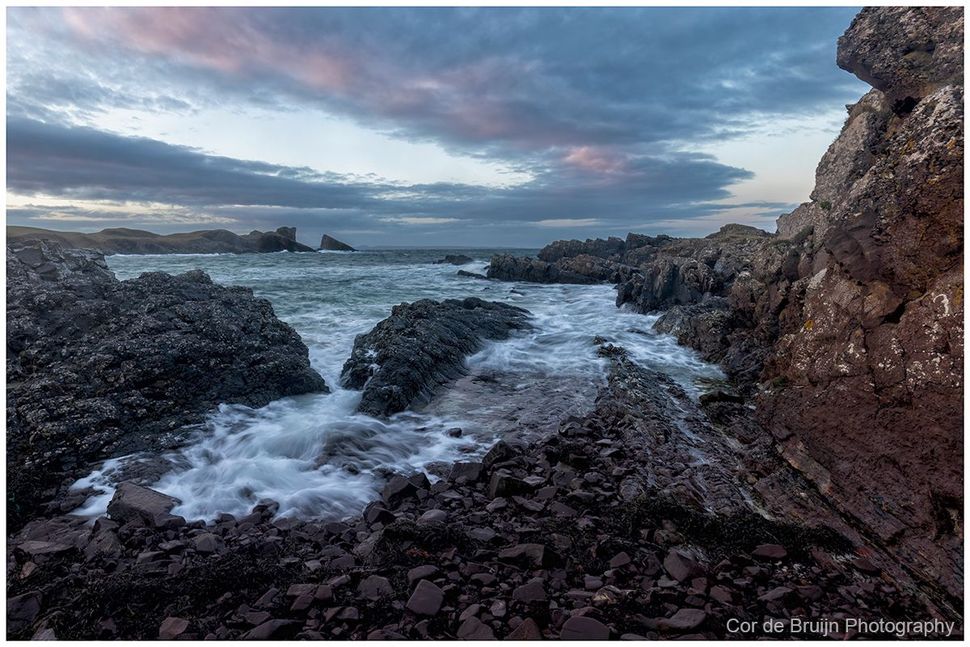 Rocky coastline with surf washing over dark stones under a dramatic, cloudy twilight sky.