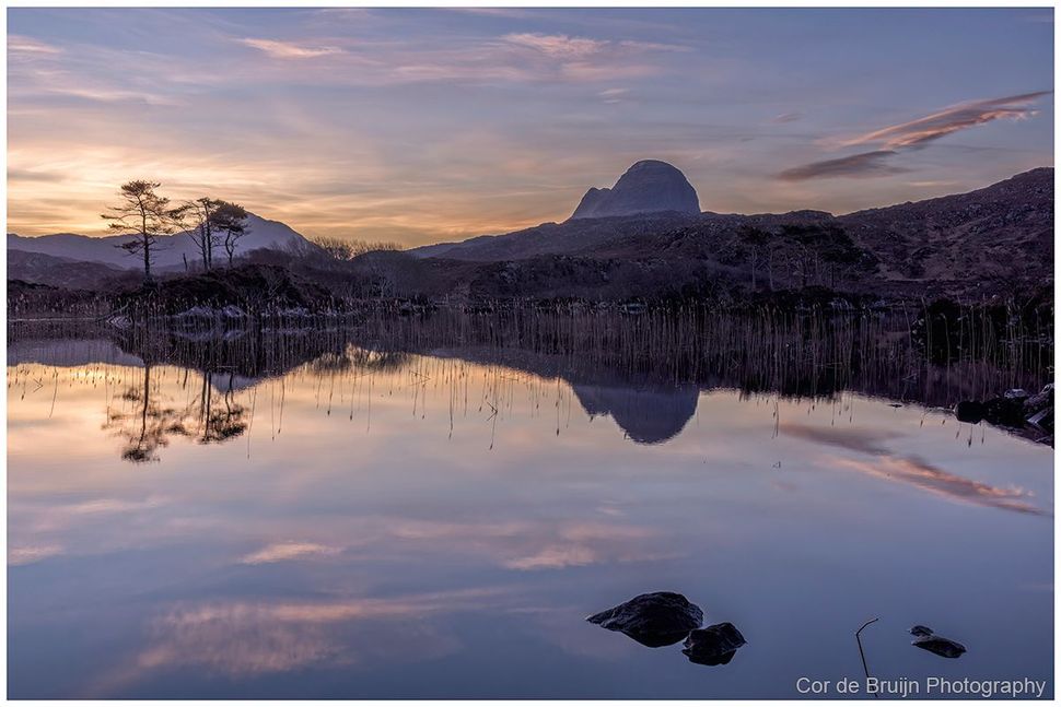 A mountain peak reflects in a calm lake at sunrise, with soft pink and blue hues in the sky and dark trees in the distance.