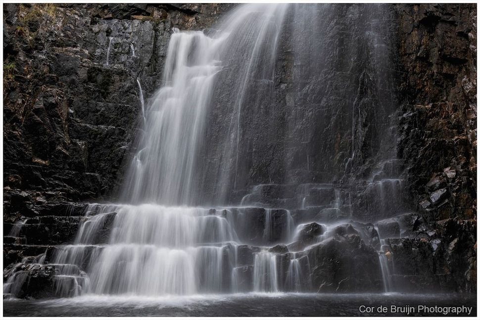 A tiered waterfall cascades down dark, textured rock cliffs in a serene, natural setting.