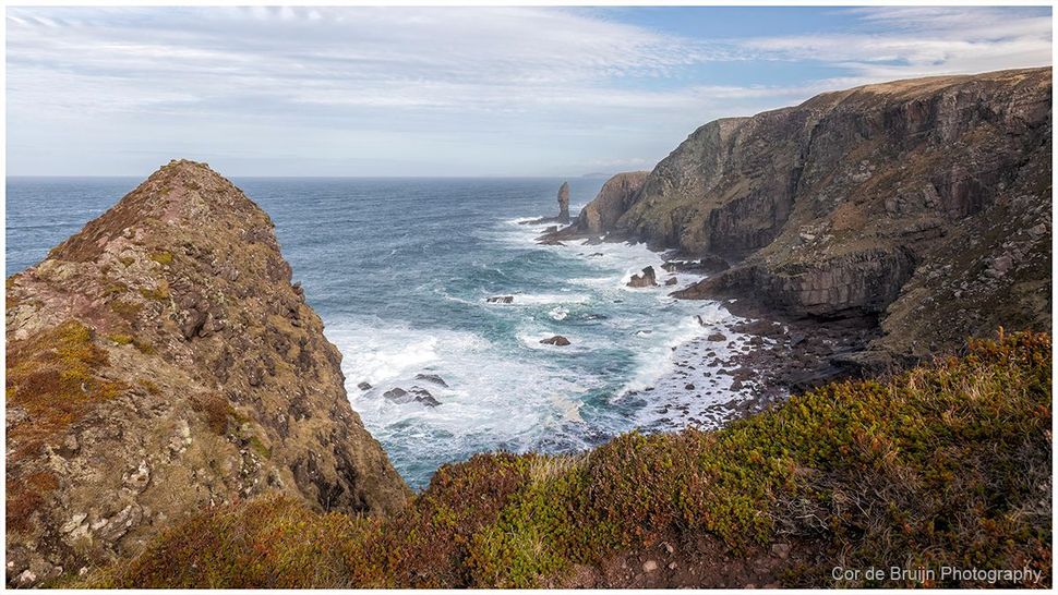 A rugged rocky cliff overlooks a churning blue ocean with white sea foam, under a cloudy sky.