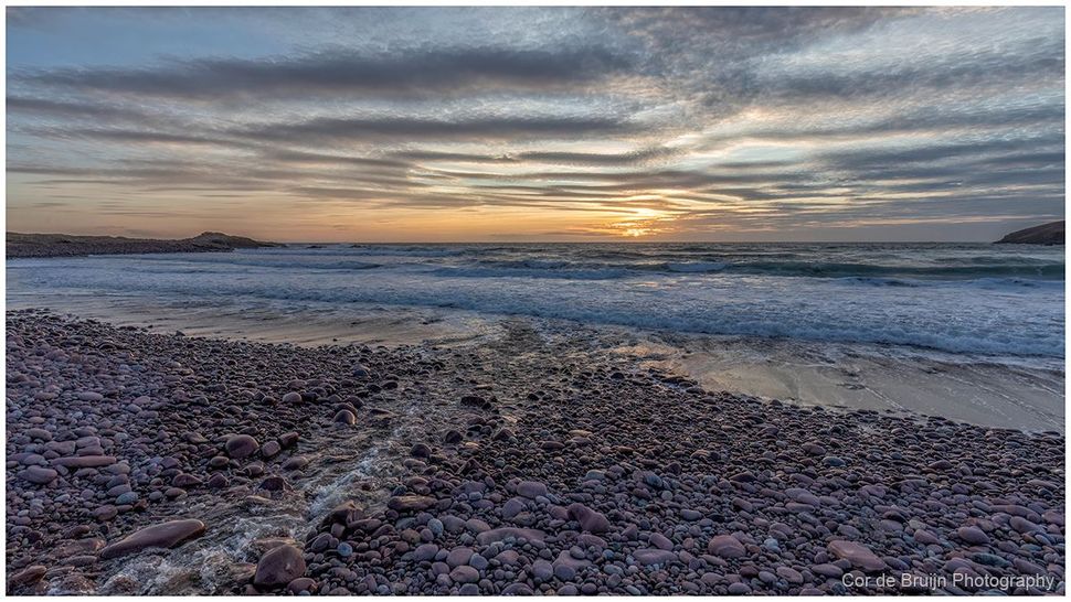A pebbled beach at sunset, with gentle waves rolling toward the shore under a dramatic, cloud-filled sky.