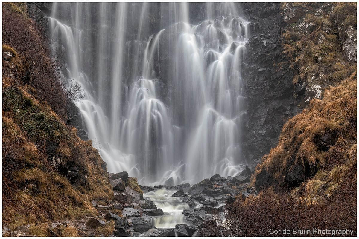 A multi-tiered waterfall flows down a dark, rocky cliff surrounded by brown, dry vegetation.