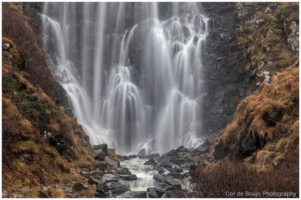 A multi-tiered waterfall flows down a dark, rocky cliff surrounded by brown, dry vegetation.