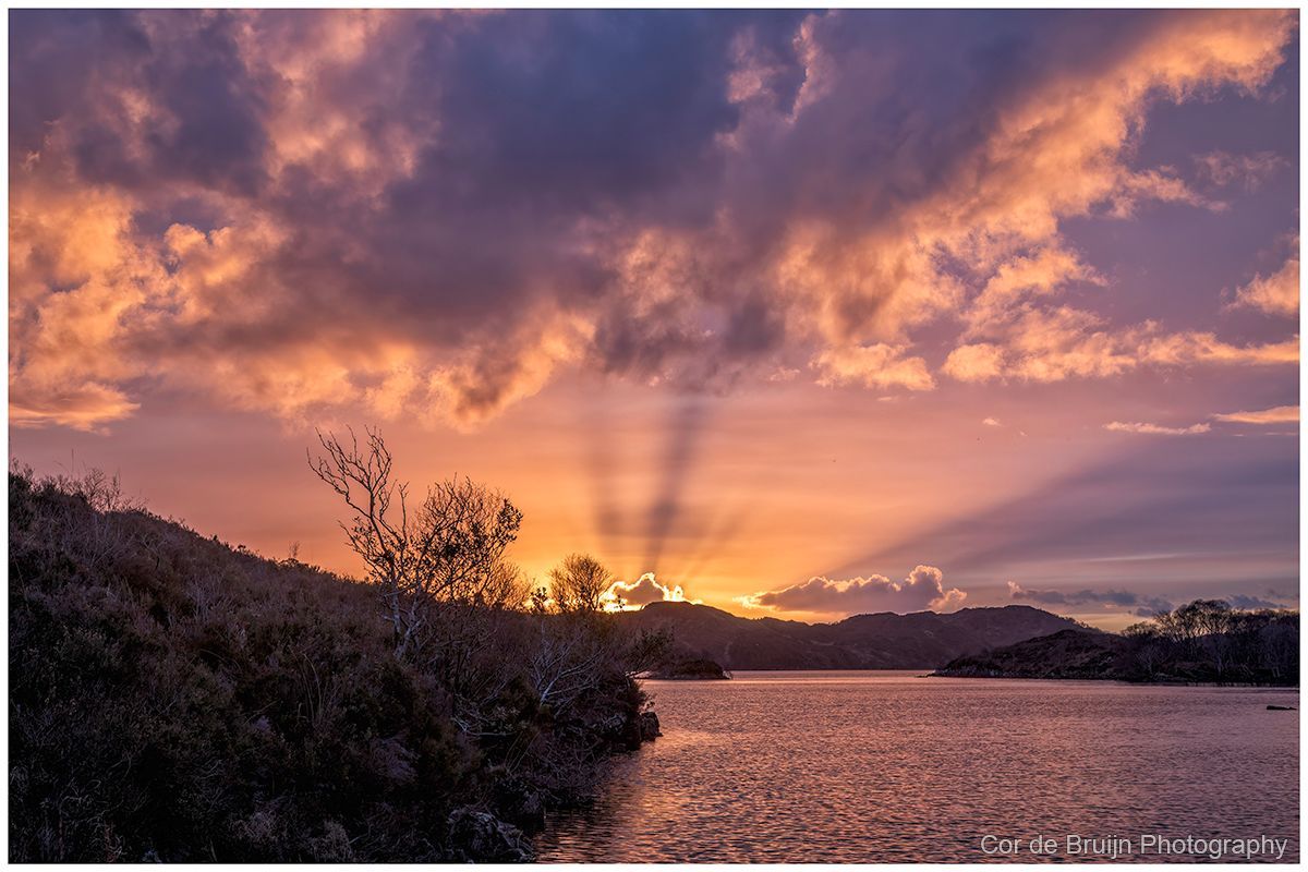 Sunset over a lake with pink-orange clouds and silhouetted trees and hills