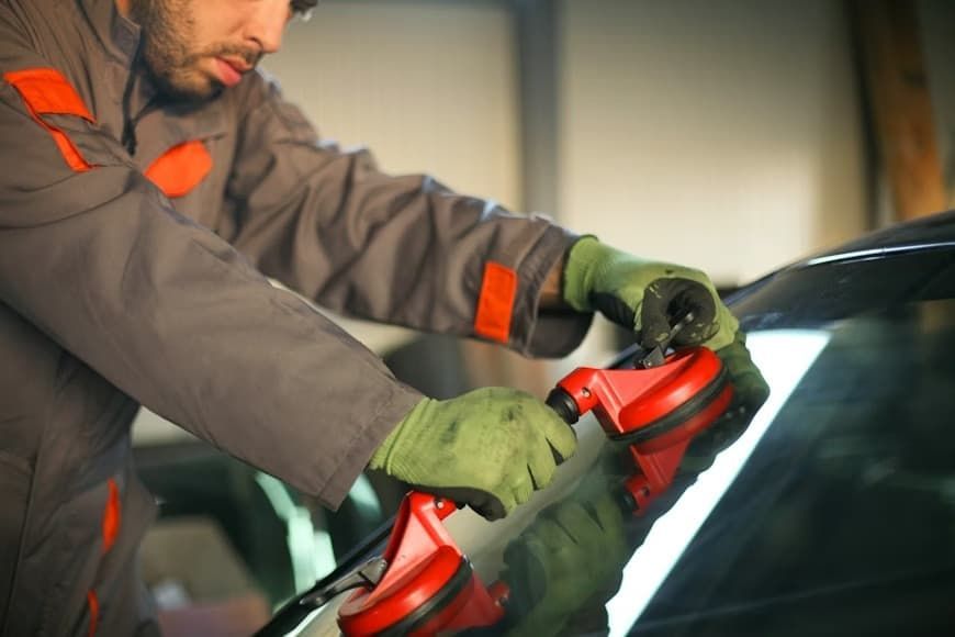 A mechanic in gray coveralls uses dual red suction cup handles to lift or position a car windshield.