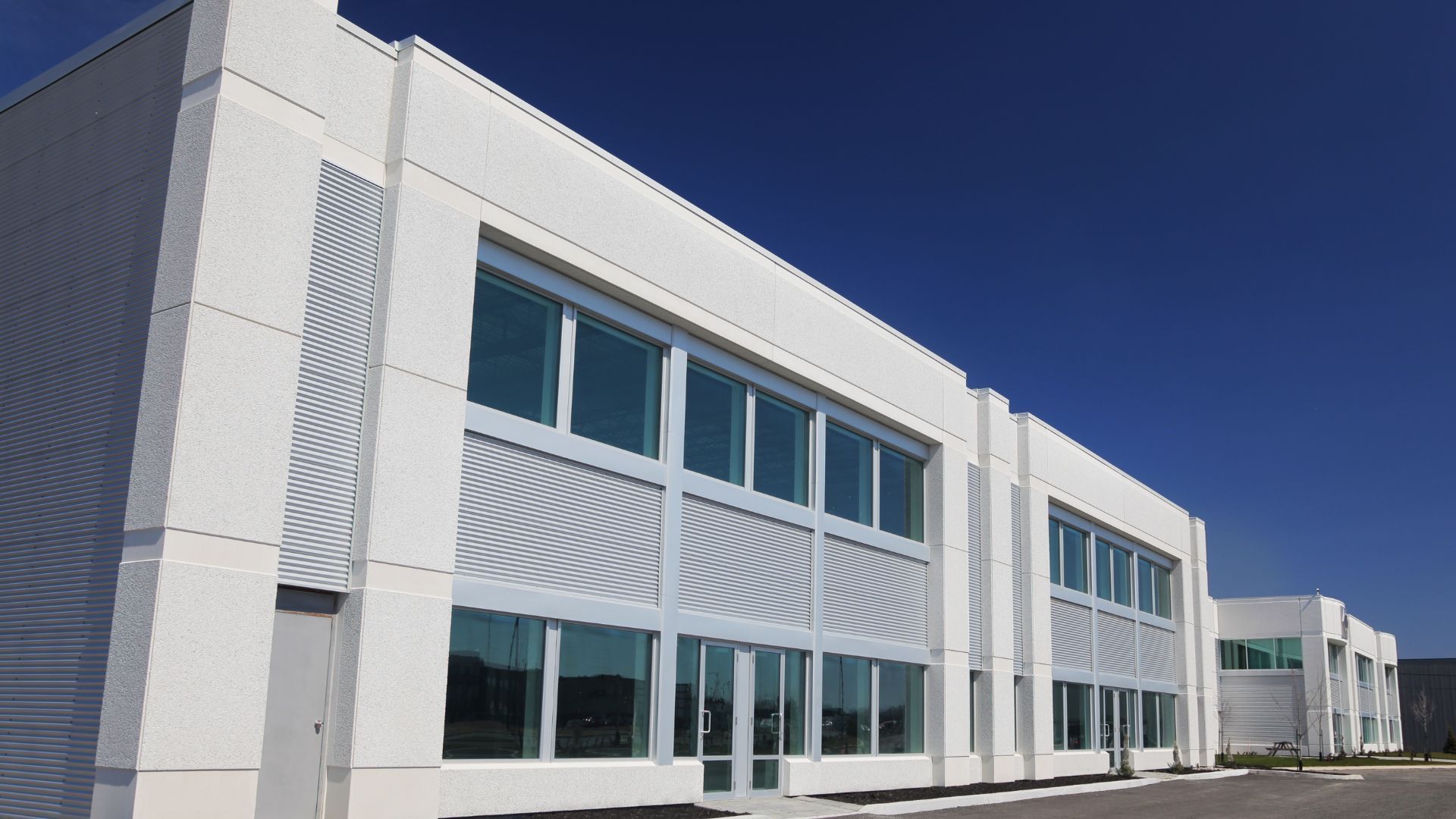 Modern white, two-story commercial building with rectangular windows and metal paneling under a clear blue sky.