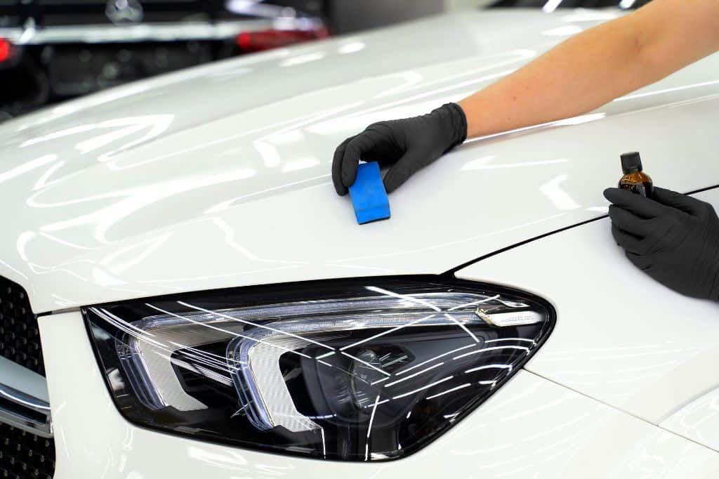 A person wearing black gloves applies a ceramic coating to a white car hood using a small blue applicator block.