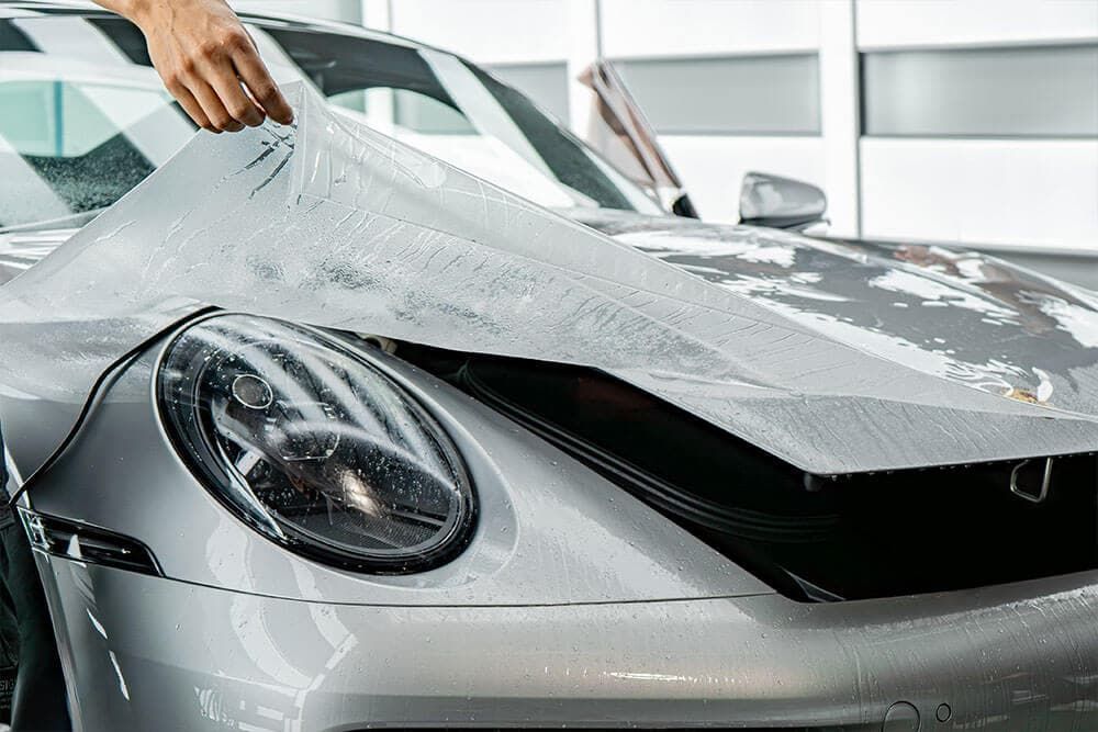 A hand peels a clear protective film off the hood of a silver sports car.