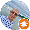 A man wearing sunglasses is taking a selfie in front of a body of water.