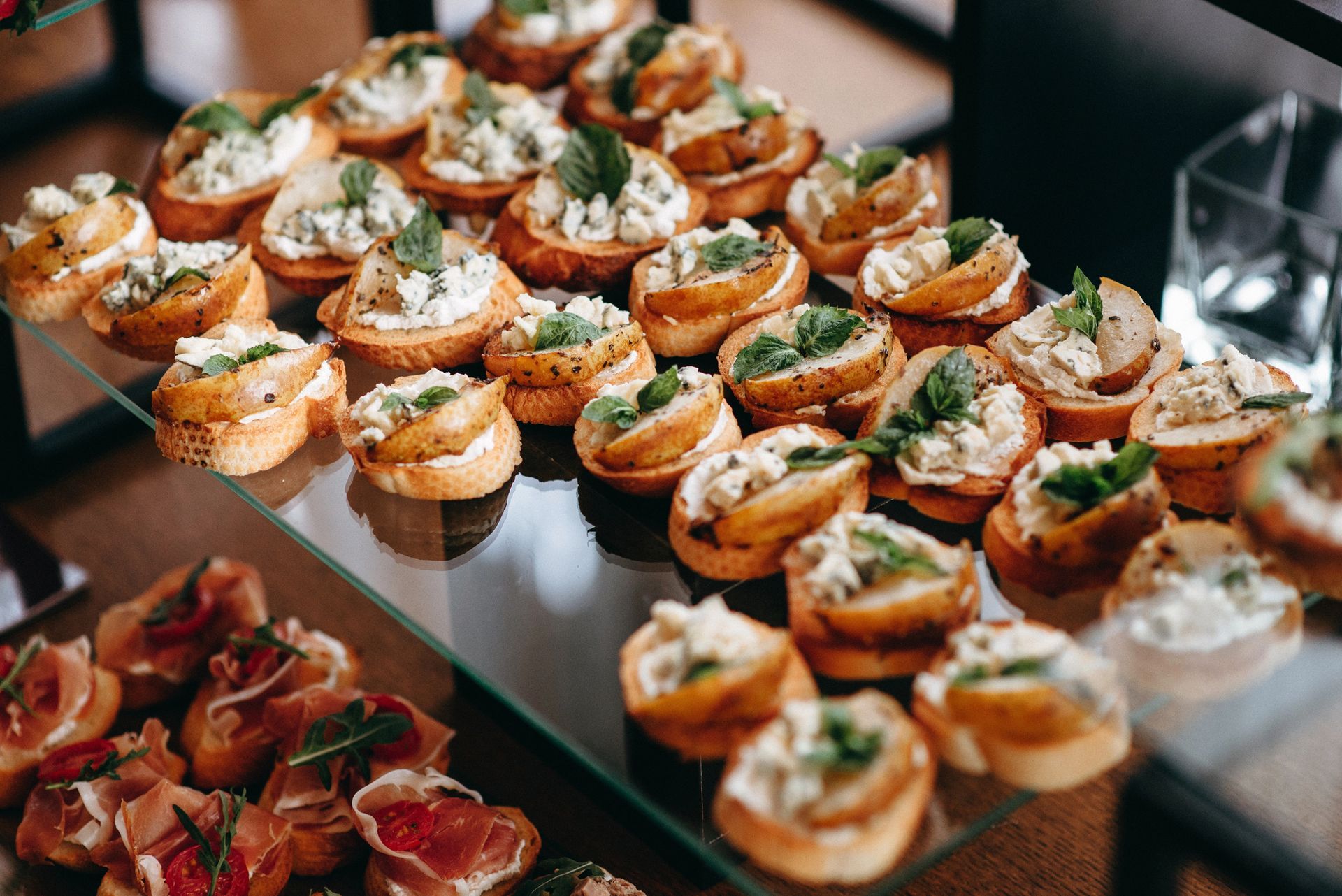 Un'alzata a più livelli in vetro, piena di vari antipasti, tra cui bruschette con formaggio e guarnizioni di erbe aromatiche.